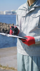 クリップボードを持つ作業服の男性・海と工業地帯│建設業・製造業・プラント　イメージ