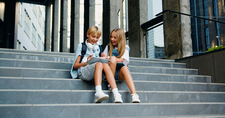Happy schoolchildren sittingding near school building on the stairs, having fun, talking about...