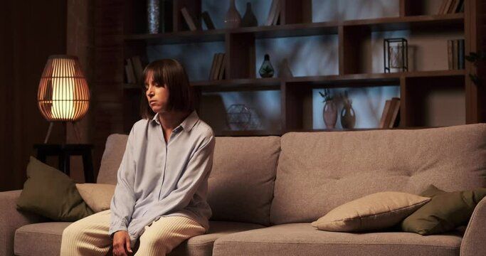 Weary woman lies down on the sofa in the living room. Her body sinks into the cushions, seeking solace and rest. The weight of the day reflects in closed eyes and relaxed posture.