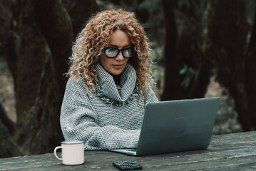 Curly long hair blonde adult woman use laptop computer outdoors sitting in the woods - concept of...