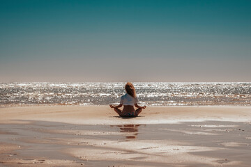 Inner balance life concept with woman sitting at the beach doing yoga meditation relax position viewed from back. Blue sky and nature sand coast lscenic place. Freedom and happiness concept