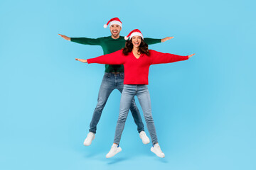 Joyful young couple in Santa hats jumping spreading arms, studio