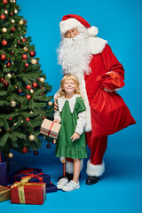 Santa Claus holding sack bag near happy girl with prosthetic leg next to Christmas tree on blue