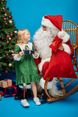 happy girl with prosthetic leg holding present near Santa Claus next to Christmas tree on blue