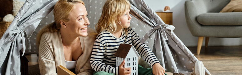 dreamy girl and her cheerful mother looking away while reading book and sitting in play tent, banner