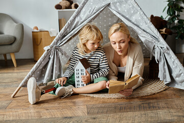 cute girl with prosthetic leg and blonde mother reading book while sitting in play tent at home