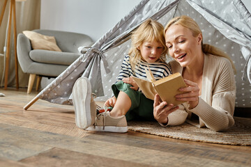 cute girl with prosthetic leg and blonde mother reading book and sitting in play tent at home