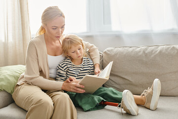 blonde mother reading book to her child with prosthetic leg while sitting together in living room