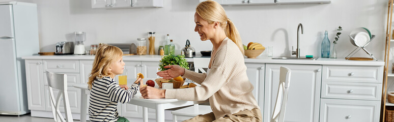 little girl having breakfast with happy blonde mother in modern kitchen, daily life banner