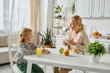 cheerful blonde mother looking at her cute daughter with prosthetic leg having breakfast in kitchen