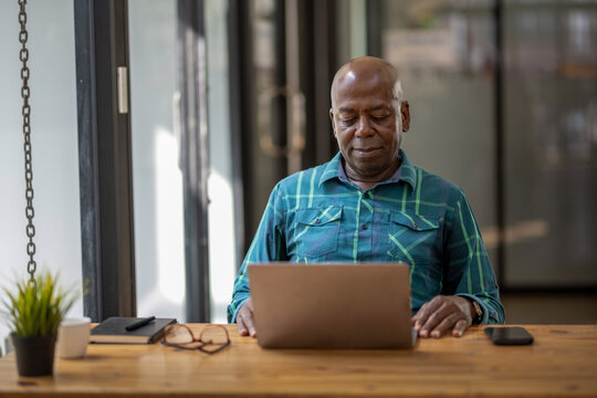 A black senior man in casual clothes is focusing on a laptop screen, concentrating and thinking seriously. - Powered by Adobe
