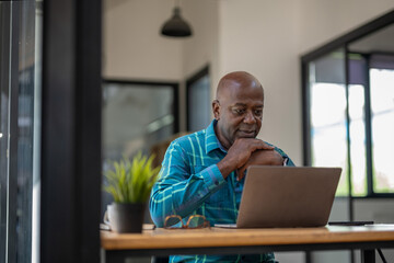 A black senior man in casual clothes is focusing on a laptop screen, concentrating and thinking...