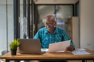 Fototapeta na wymiar A senior black man in casual clothes is busy at work but happy with the work and paperwork in front of him.