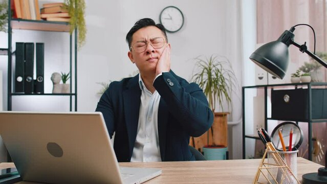Dental problems. Asian businessman working at office touching cheek, closing eyes with expression of terrible suffer from painful toothache, sensitive teeth, cavities. Male Chinese in formal suit