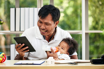 Young Asian father looking digital tablet smiling and treat baby boy feeding milk and working from home office