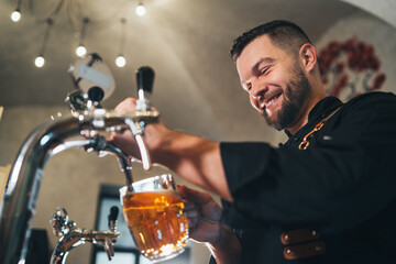 Smiling stylish bearded barman dressed black uniform with an apron tapping fresh lager beer into glass mug at bar counter. Successful people, beer consumption, beverages industry concept image - Powered by Adobe