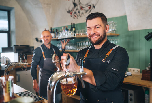 Stylish bearded barman dressed black uniform smiling at camera, beer tapping at bar counter and waiter with tray. Successful people teamwork, friendship, brewing and restaurant industry concept image