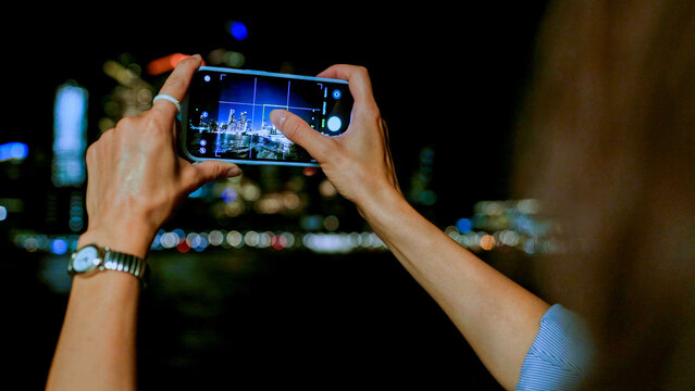 Over the shoulder view of beautiful woman's hands holding smartphone and taking pictures of picturesque city at night. Young attractive girl tapping on screen to focus. Camera concept.