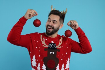 Happy young man in Christmas sweater and reindeer headband holding festive baubles on light blue background - obrazy, fototapety, plakaty