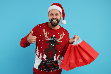 Happy young man in Christmas sweater and Santa hat with shopping bags on light blue background