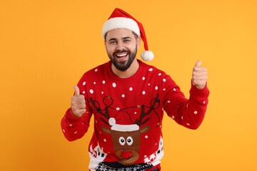 Happy young man in Christmas sweater and Santa hat showing thumbs up on orange background