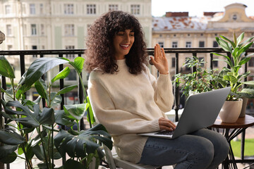 Beautiful young woman using laptop surrounded by green houseplants on balcony