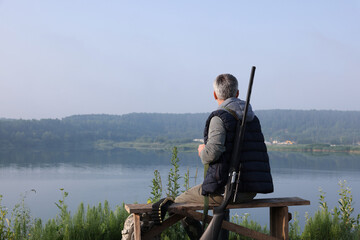 Man with hunting rifle sitting on wooden bench near lake outdoors, back view. Space for text
