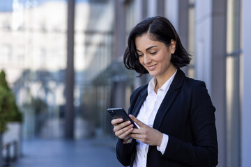 Fototapeta premium A young beautiful business woman is walking outside the office building, a woman in business clothes is smiling, using an application on the phone, browsing the Internet, typing a message