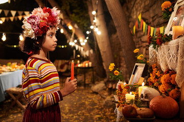 Little girl in traditional clothes standing with candle at altar in honour of dead people