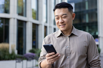 A smiling young man businessman Asian stands on the street of the city, holds a phone in his hand,...