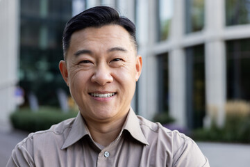 Close-up portrait of a smiling young Asian man in a brown shirt standing on a city street and...