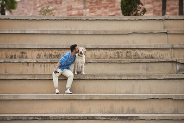 Young Hispanic man, sitting on big cement stairs giving a kiss to his dog in loving and tender attitude. Concept, dogs, pets, animals, friends.