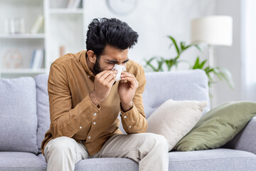 Fototapeta premium Sick man sitting at home on sofa in living room, sneezing has allergy and runny nose, Indian man with napkin has a cold.