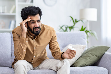 Fototapeta premium Sick man sitting at home on sofa in living room, sneezing has allergy and runny nose, Indian man with napkin has a cold.