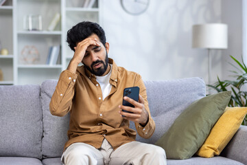 Sad unhappy and disappointed man sitting at home on sofa in living room, Indian man received...