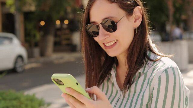 Close up video of happy young woman in causal typing a message on phone outdoors in city.