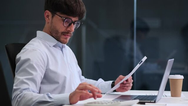 Inside a busy office space, a dedicated businessman seamlessly juggles tasks, managing both papers and a laptop. His focused demeanor illustrates multitasking abilities, demonstrating efficiency.