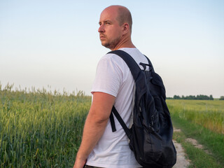 a man stands half-turned on a path in a field with a black backpack with his back to the camera. Evening landscape, the concept of travel, recreation, hiking, new discoveries