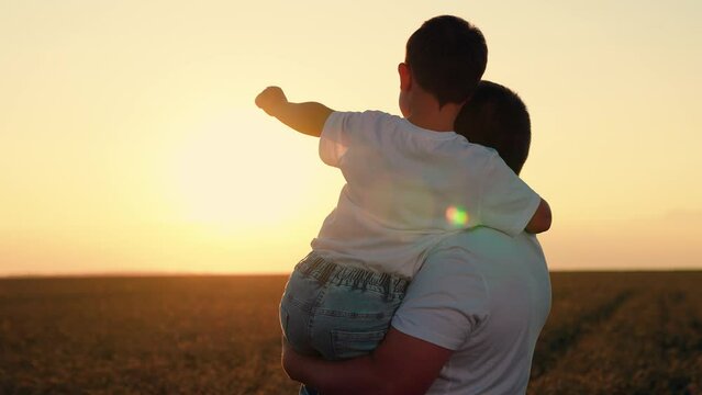 Dad holds son in arms and boy points at setting sun in field