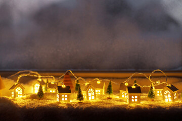 Cozy christmas miniature village. Stylish cute little glowing houses and christmas trees on soft snow blanket with lights in evening room. Atmospheric winter village still life. Merry Christmas!