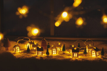 Atmospheric miniature winter village. Stylish cute little glowing houses and christmas trees on soft snow blanket with golden lights bokeh in evening room. Christmas background. Happy Holidays!