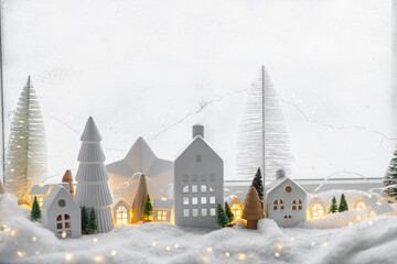 Cozy christmas miniature village. Stylish cute little ceramic houses and wooden trees on soft snow blanket with glowing lights in room. Atmospheric winter village still life. Merry Christmas!