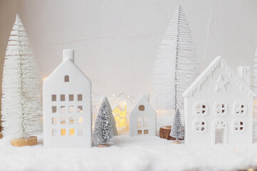 Atmospheric miniature white christmas village. Stylish little christmas houses and trees on snow blanket with glowing lights. Christmas modern winter village, holiday banner. Happy Holidays!