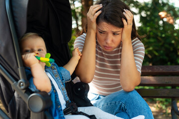 Tired young mother sits on a bench upset next to a baby stroller