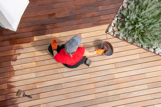 Spring deck maintenance concept. Coating exterior wooden flooring of a terrace with brown natural oil protection. Overhead view of worker on hardwood decking