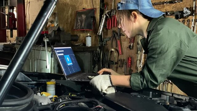 Young woman in coveralls works at a car service station prepares for computerized car diagnostics indoors. Female mechanic using laptop, software app to test scan components systems, check issues