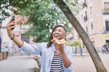 attractive man with braids holding a slice of pizza in his hand while taking a photo with his smart phone on the street