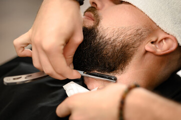 A barber shaves the neck of a bearded customer with dangerous razor. Shaving the contour of the beard for the correct shape.