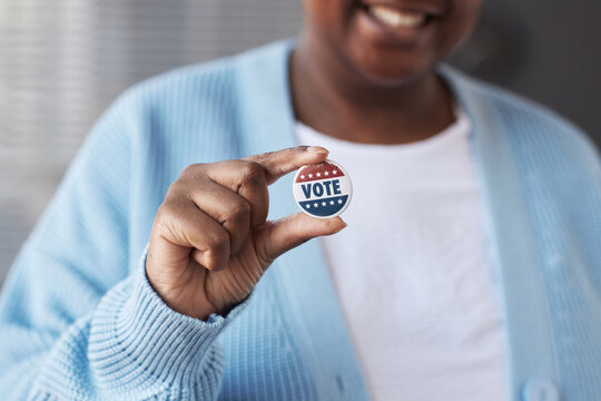 Focus on hand of young African American woman in casualwear holding small round vote badge while standing in front of camera