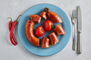 Fried pork sausage cut into portions and grilled tomatoes on a blue ceramic plate with cutlery and...